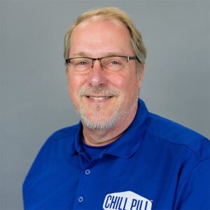 Smiling man with glasses and a short beard wearing a blue polo shirt that says "CHILL PILL" on the chest, posed in front of a plain gray background.