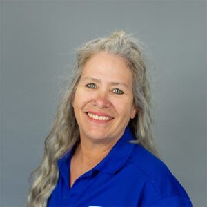 A woman with long gray hair and a blue collared shirt smiles at the camera against a plain gray background.