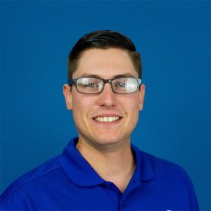 A man with short dark hair and glasses, wearing a blue collared shirt, smiles in front of a plain blue background.