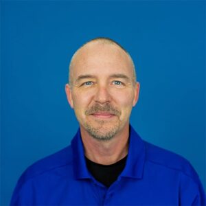 A middle-aged man with short hair and a trimmed beard, wearing a blue collared shirt, smiles in front of a solid blue background.