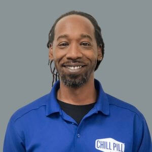 A man with a beard and dreadlocks smiles at the camera, wearing a blue polo shirt with a "Chill Pill" logo, against a plain gray background.