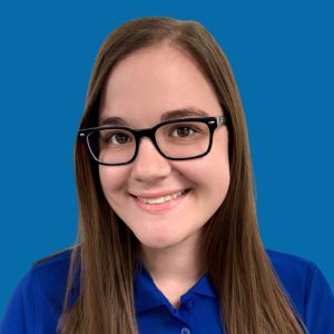 A young woman with long brown hair and glasses smiles at the camera, wearing a blue collared shirt against a solid blue background.