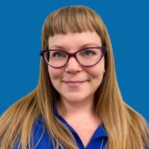 A woman with straight, light brown hair, bangs, and purple glasses is smiling at the camera against a solid blue background.