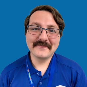 A man with glasses and a mustache, wearing a blue collared shirt and a lanyard, stands in front of a solid blue background.