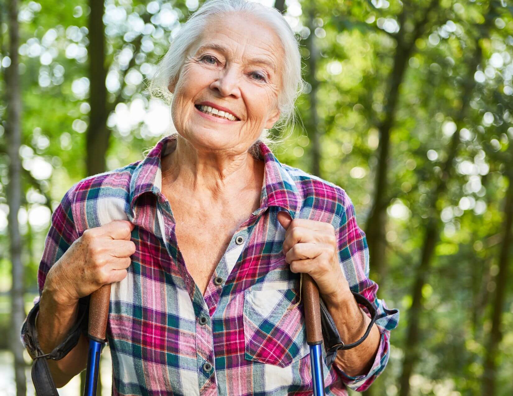 Older woman in a plaid shirt stands outdoors in a forested area, smiling and holding hiking poles.