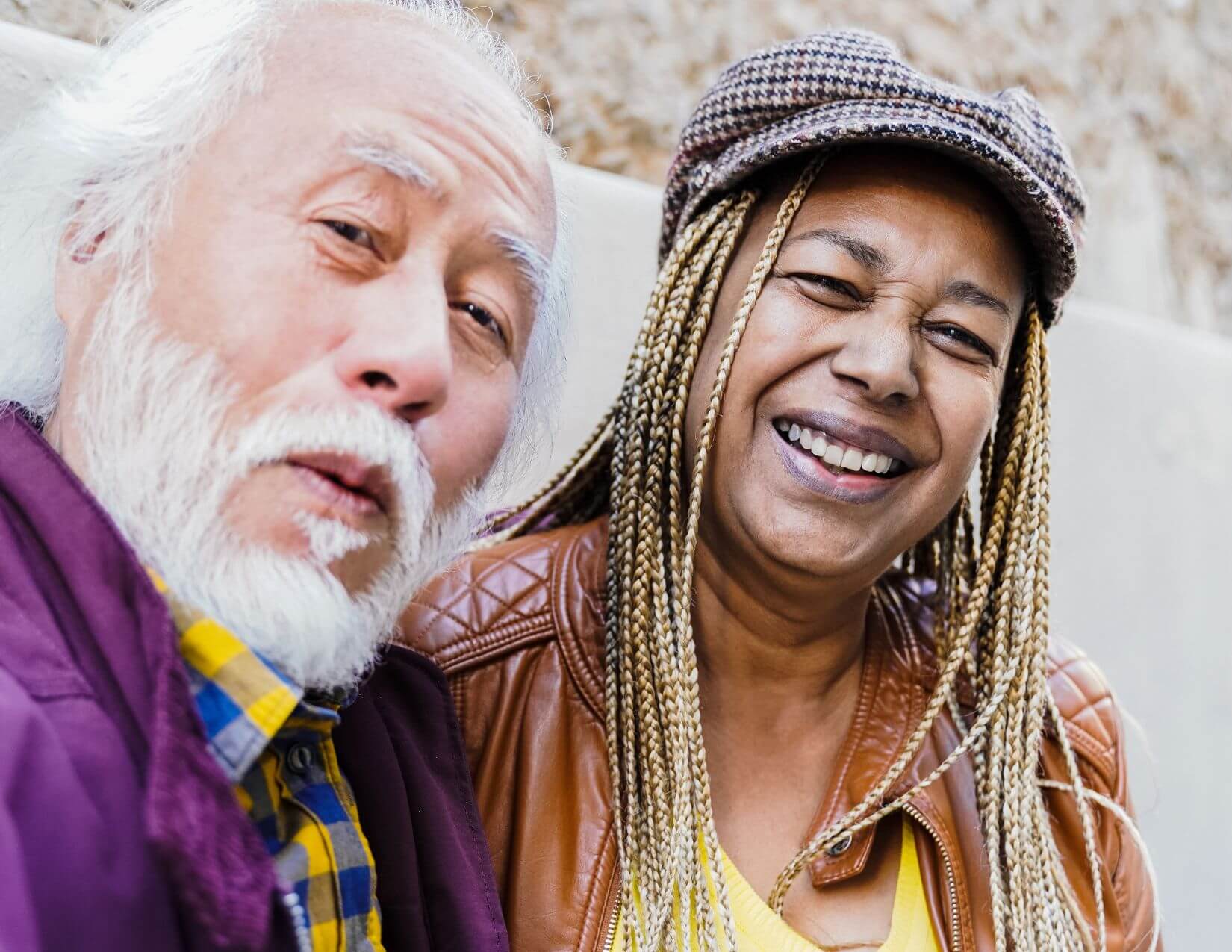 An older man with white hair and beard and a woman with braided hair and a cap are sitting together, smiling at the camera.