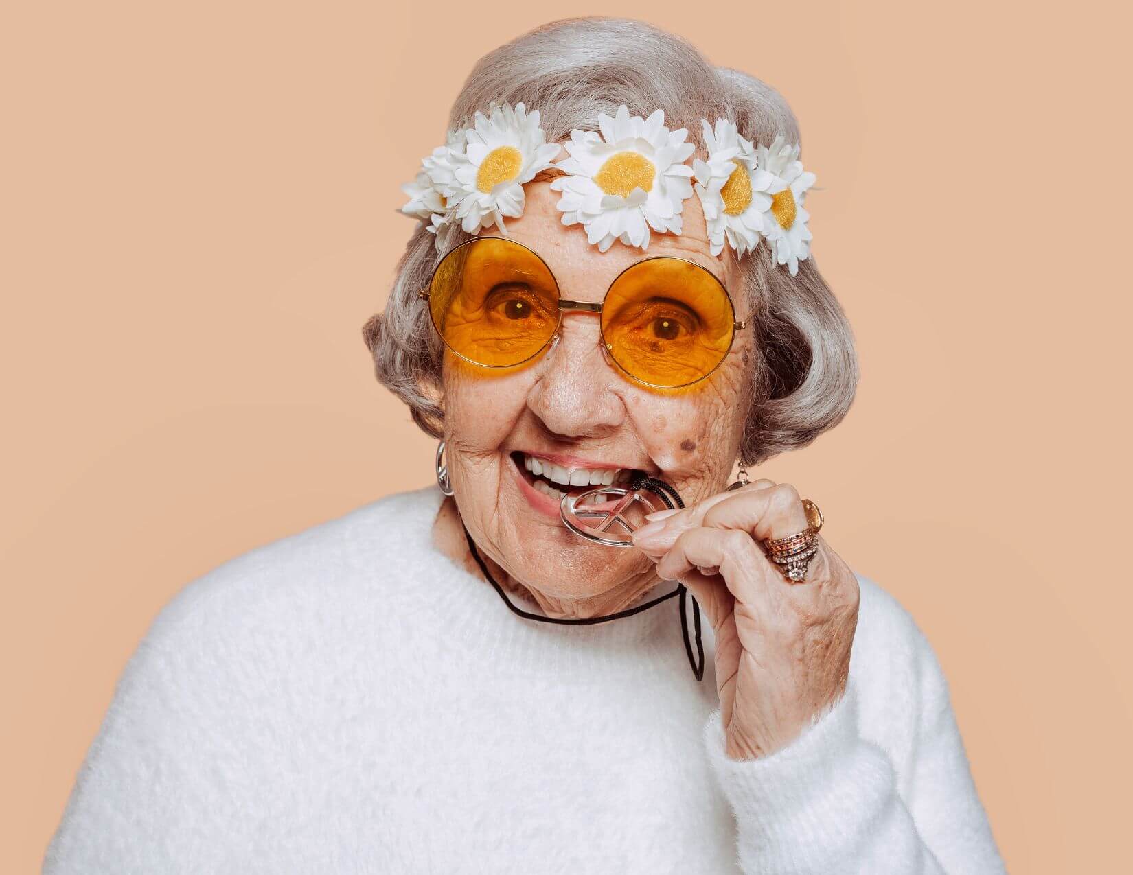 Elderly woman with gray hair, wearing yellow-tinted glasses, a white sweater, and a daisy flower crown, smiling and playfully biting her eyeglass frames against a peach background.