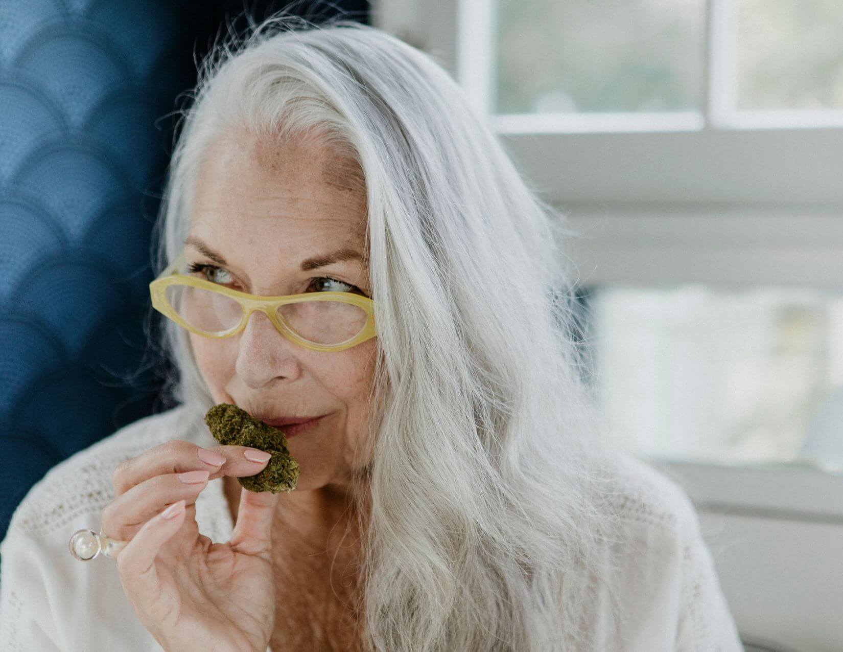 Older woman with long gray hair and yellow glasses holds and smells a green edible, possibly a cannabis-infused treat, in a light-filled room.