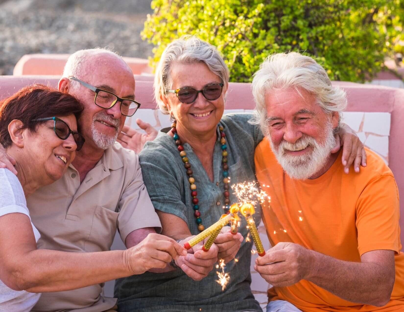 Four older adults sit closely together outdoors, smiling and holding sparklers, with greenery and a pink wall in the background.