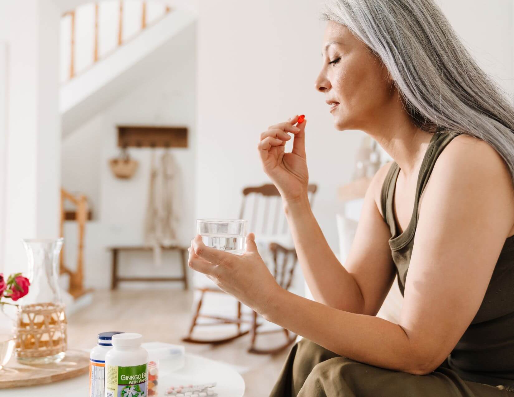 A woman with long gray hair holds a pill and a glass of water, sitting indoors near a table with medicine bottles and a vase of flowers.