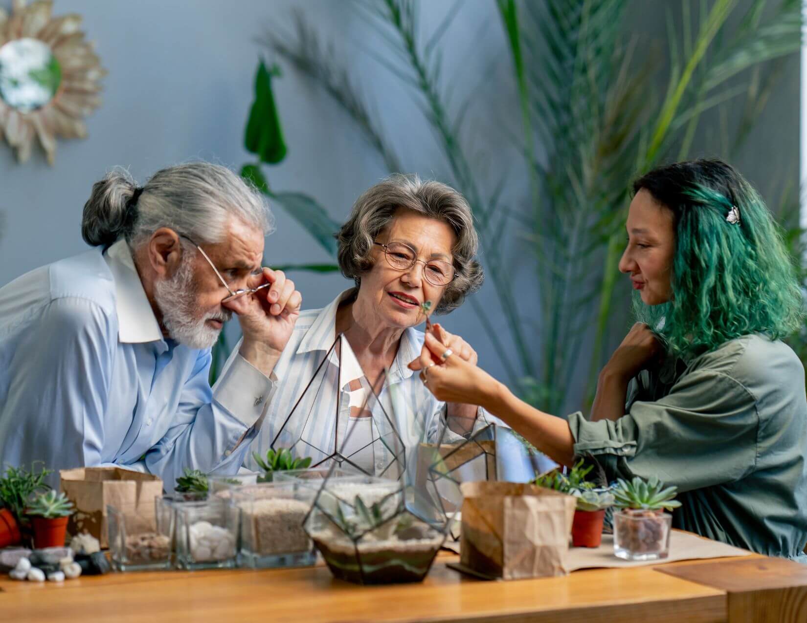 Three people sit at a table working together to arrange succulents and other plants in terrariums, surrounded by pots, soil, and greenery.