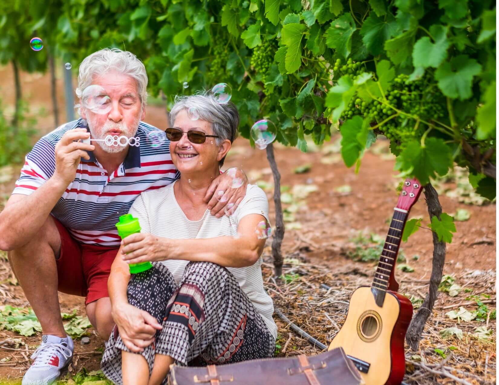 An older couple sits outdoors near grapevines; the man blows soap bubbles while the woman smiles. A guitar and bag are on the ground beside them.