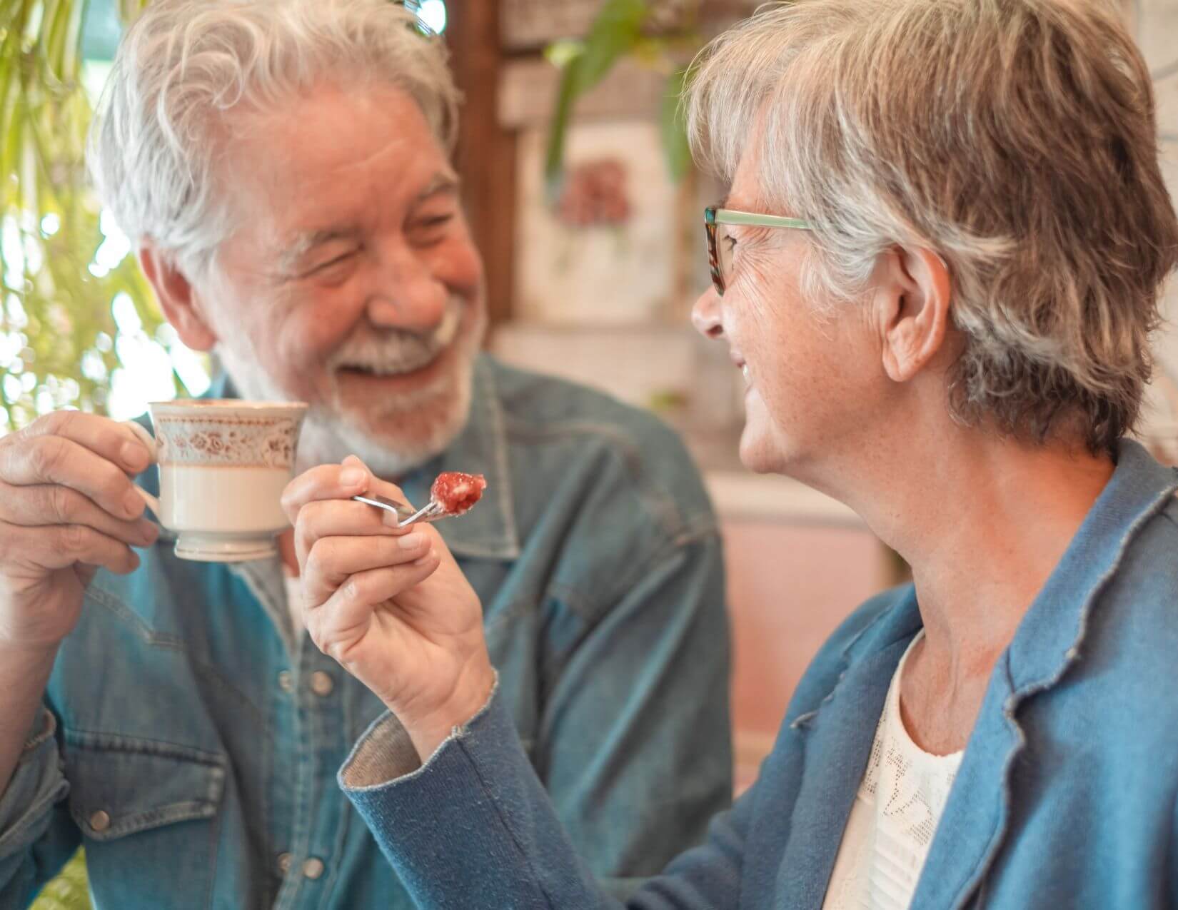 An older man and woman sit together, smiling. The man holds a teacup while the woman offers him a spoonful of food. Both are wearing glasses and casual clothing.