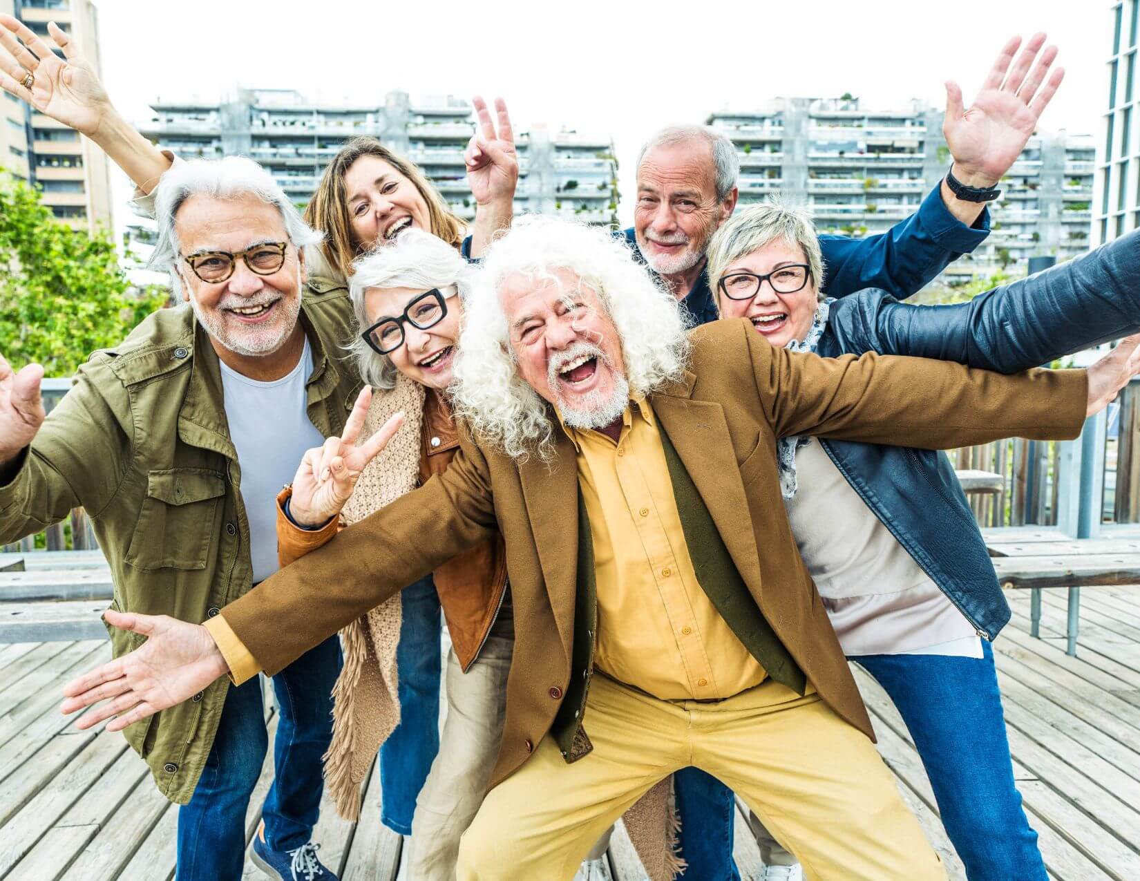 Six older adults stand outdoors on a wooden deck, smiling and posing energetically for the camera, with buildings visible in the background.
