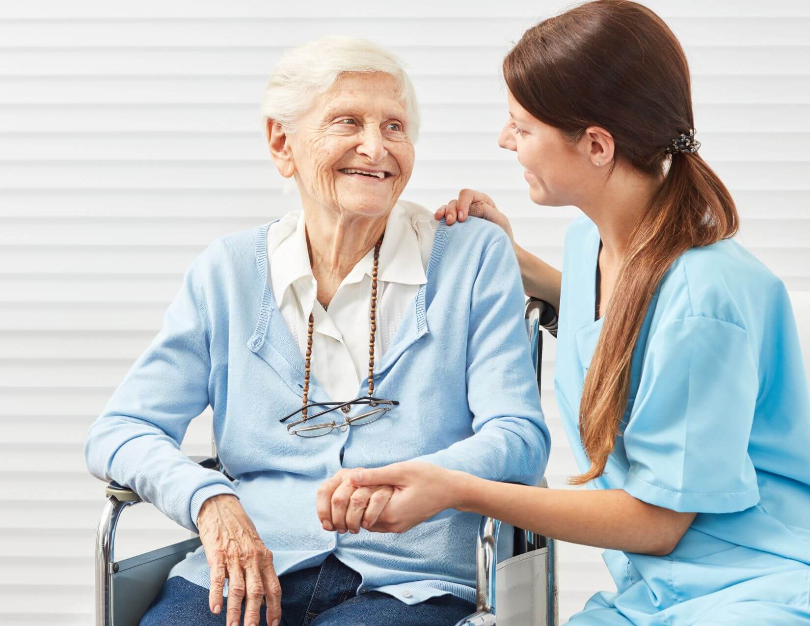 A young caregiver in blue scrubs holds the hand of an elderly woman in a wheelchair. Both are smiling and making eye contact in a bright indoor setting.