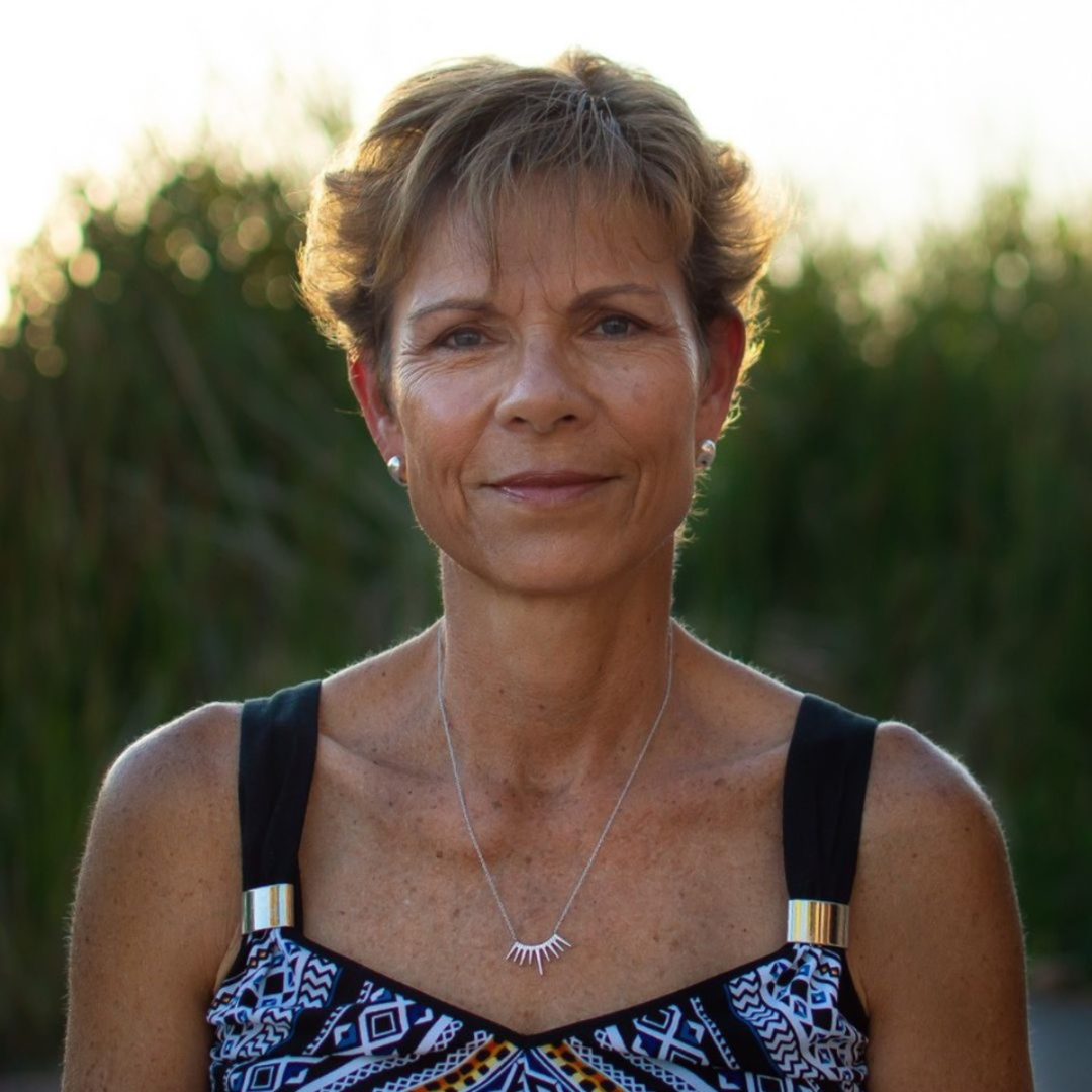 A middle-aged woman with short hair wearing a patterned dress and a necklace stands outdoors with greenery in the background.